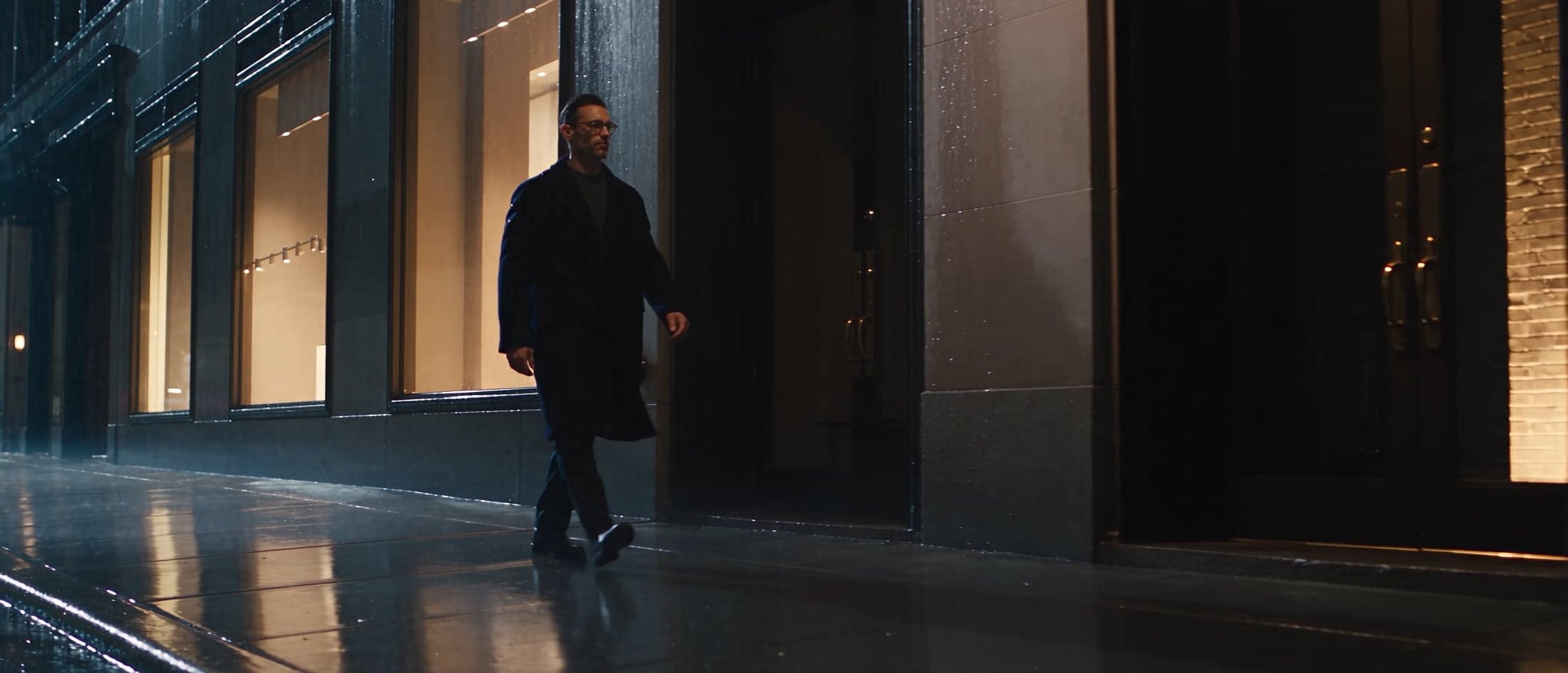 A man in glasses and a dark coat walks past illuminated storefront windows on a rain-slicked street at night.